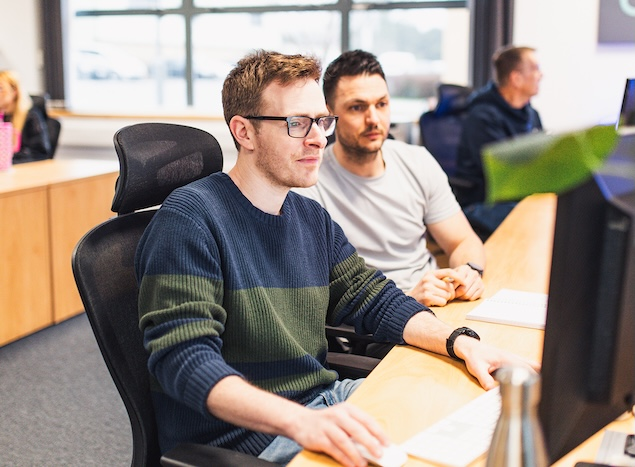 Two male Bluestorm colleagues, one wearing glasses, focused at a desktop in a bright open-plan office.